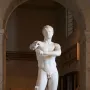 Apoxyomenos, a white marble nude male statue, shown under a museum archway scraping his left forearm.