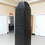 Black Obelisk with stepped top and carved relief bands on a wooden base in a museum interior.
