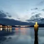 Havmannen sculpture standing in calm water at twilight, with its reflection visible and an illuminated industrial area in the background.