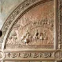 Close-up of a bronze gate relief showing horsemen in a historical landscape with Latin inscription and decorative scrollwork.