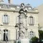 Gavarni Fountain with a stone column topped by a figure holding a book, with relief sculpture and sculpted heads at the base.