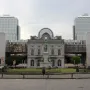 The John Cockerill Monument stands on a pedestal in a circular grassy area, with a historic building and modern glass towers behind it.