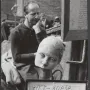 A sculpted female head resting on a metal pitcher, with a man in the background and a framed handwritten sign at right.
