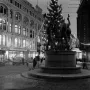 The Three Smiths statue with three muscular figures holding hammers around an anvil in a city square, with a lit Christmas tree behind it.