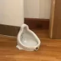 A white glazed ceramic urinal sits on a polished wooden floor, with dark dots visible inside and a circular drain opening near the base.
