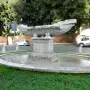 Fontana della Navicella, a weathered stone boat-shaped fountain in an oval basin surrounded by grass and trees.