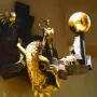The Defender of Time sculpture with a golden figure, clock, rooster, and creature mounted on a wall.