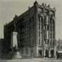 The Brigham Young Monument with a bronze figure atop a tall stone pedestal and relief sculptures at the base, with a white building and bare trees behind it.