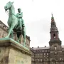 Christian IX equestrian statue on a tall stone pedestal in an open square, with ornate buildings and a slender tower behind it.