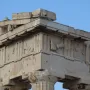 Weathered marble corner of the Parthenon pediments with fluted columns, triglyphs, worn relief metopes, and a lion\'s head against a blue sky.