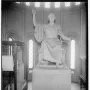 A large seated marble statue of a male figure with one arm raised and the other holding a bound object, displayed in a room with arched windows.
