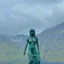 Kópakonan bronze statue standing on dark rocks by the water with misty mountains behind it.