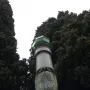 Lighthouse of Lierna, a tall stone monument with a green lantern top beside dense trees.