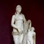 Venus Felix marble statue with a small winged child beside her, set on a inscribed plinth against a dark red background.