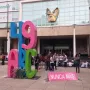Antimonumento 49 ABC stands on a paved plaza in front of a modern building, with a small bouquet of white flowers and a pink sign reading \