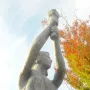 Victims of Communism Memorial statue viewed from below, with the female figure raising an object overhead against a bright sky and autumn leaves.