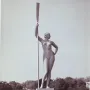 Girl with an Oar sculpture standing on a tiered pedestal in a park fountain, holding a tall oar upright.