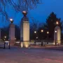 Commonwealth Memorial Gates at dusk, with a domed pavilion, four lit stone pillars topped by flames, and bare trees against a blue sky.