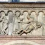 Stone relief from Scenes from Genesis showing several figures beneath small arches, including clothed people gesturing toward nude bodies and a tree at the right.