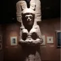 The Young Woman of Amajac sculpture displayed on a white pedestal in a dim museum hall.