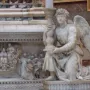 Michelangelo\'s Angel kneeling and holding an ornate candelabra beside carved marble relief.