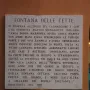 Marble plaques on a reddish-brown wall beside the Fontana delle Tette, with one plaque in uppercase text and a smaller cursive plaque below.