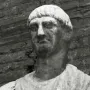 Weathered stone bust of Abbot Luigi with a serious expression, set against a dark brick background.