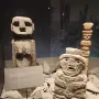 Chemamull wooden figure displayed among grey stones with smaller stone sculptures nearby.