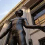 Bronze statue of Jean de Fiennes with arms outstretched, seen from below against a stone building.