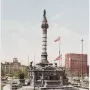 Soldiers\' and Sailors\' Monument rising from a stepped base in a landscaped park, with American flags nearby and city buildings in the background.
