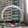 The Eye of Mexico sculpture in an urban plaza beside a tall gridded building, framed by trees and city streets.