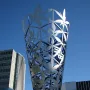 The Chalice metal sculpture rising against a blue sky in Christchurch, with modern buildings behind it.