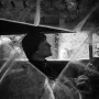 Surreal black and white scene of Rainy Taxi seen through a fractured windshield, with a mannequin in the driver\'s seat and reflections of two people in the windows.