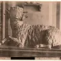 Resting bronze ram with curled horns on a long pedestal, shown against a striped and pillared wall backdrop.