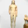 A tall marble Kouros statue of a nude male figure standing frontally with long braided hair and a missing left forearm.