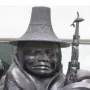 Close-up of Spirit of Haida Gwaii showing a carved figure in a woven-textured hat and patterned cloak beside a whale-topped staff.