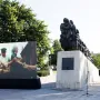 Train of Pain monument on a light stone base in a paved square, with a large historical display screen beside it.
