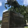 Lower section of the Simón Bolívar equestrian statue on a dark stone pedestal with gold inscriptions, framed by trees and a cloudy blue sky.